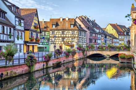 Quartier de la Petite France à Strasbourg avec maisons à colombages colorées bordant un canal, pont fleuri et reflets des façades dans l’eau.