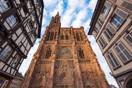 Vue en contre-plongée de la cathédrale Notre-Dame de Strasbourg, avec sa façade gothique détaillée et sa flèche élancée, encadrée par des bâtiments traditionnels à colombages et volets blancs sous un ciel partiellement nuageux.